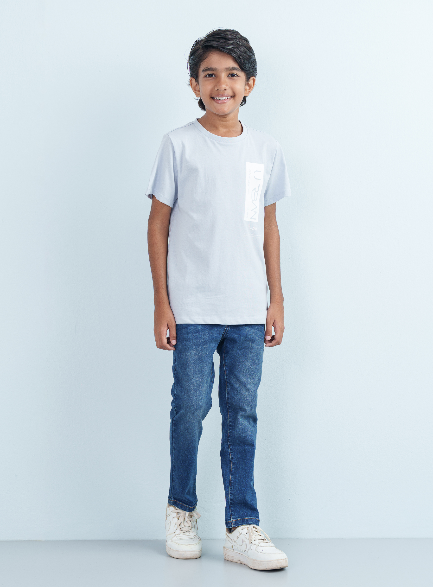 Full-length studio shot of a smiling young boy wearing a light gray cotton T Shirt Half with an urban chest graphic paired with blue jeans.