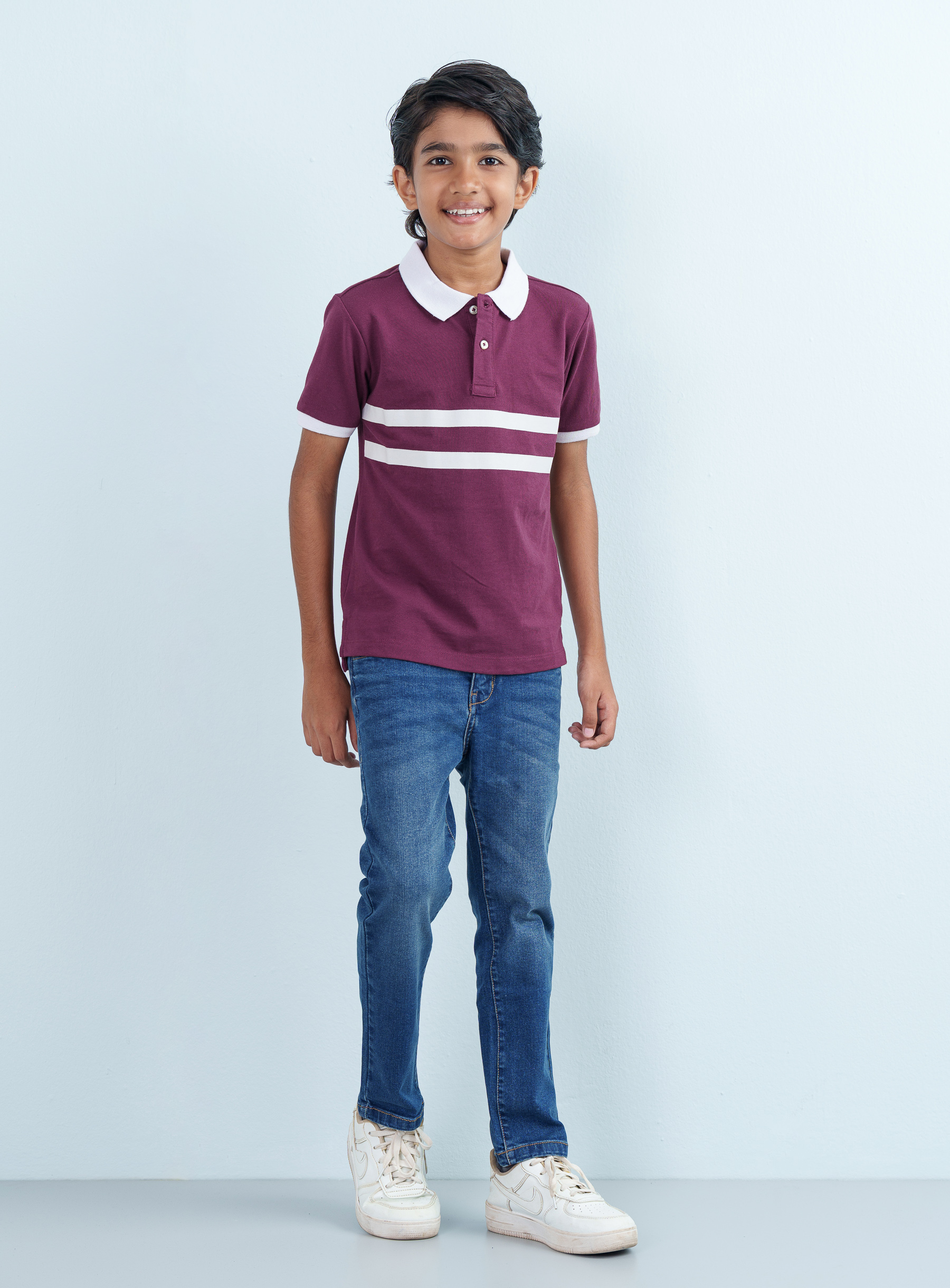 Full-length front shot of a smiling young boy modeling a maroon Polo Half with dual white stripes paired with classic blue jeans.