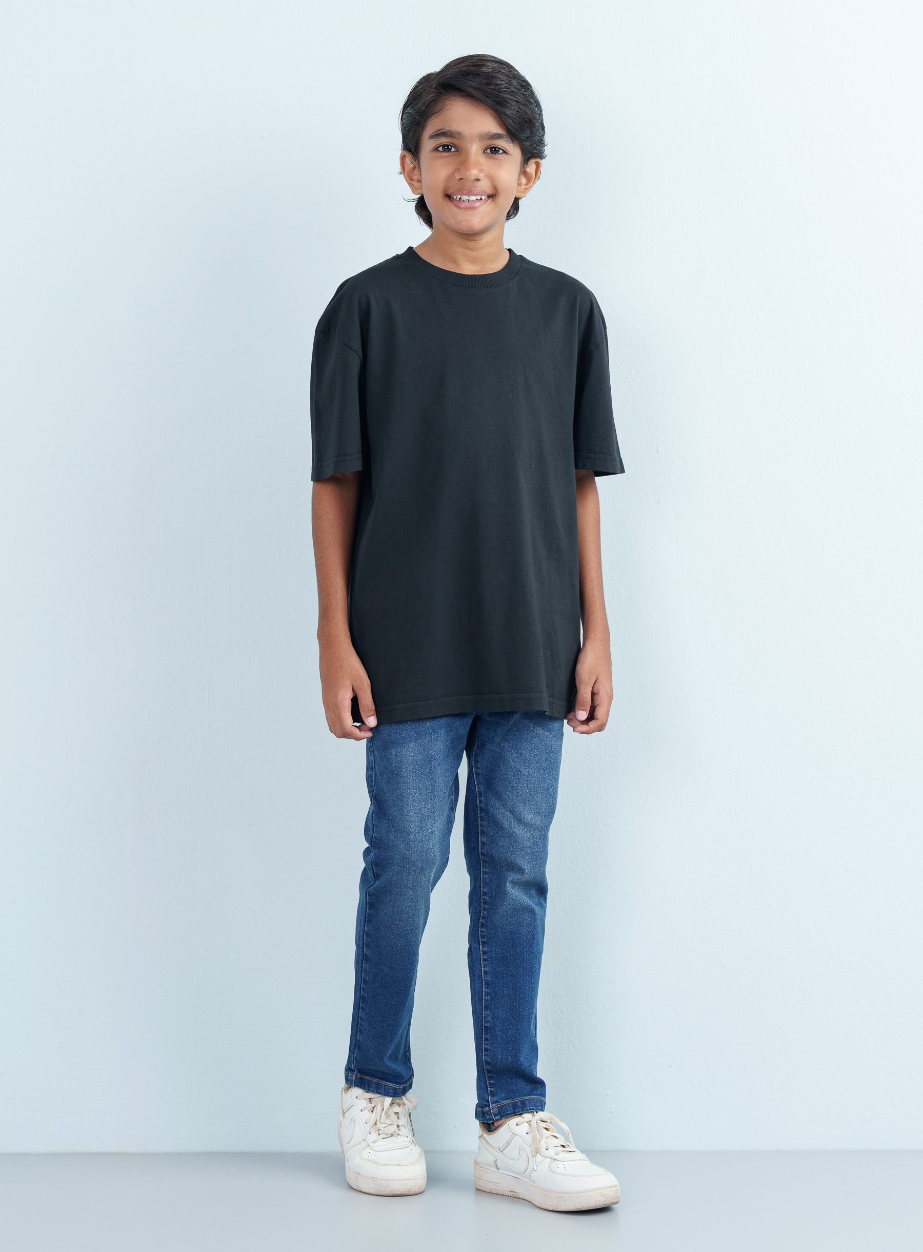 Full-length front shot of a smiling boy modeling a casual black T Shirt Half with an oversized fit paired with classic blue jeans.